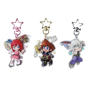 Puede incluir: Tres llaveros de acrílico con personajes estilo chibi de Kingdom Hearts. Cada dije cuelga de un broche en forma de estrella. El dije de la izquierda muestra un personaje de pelo rojo en rosa, el central un personaje de pelo castaño y el de la derecha uno de pelo blanco.
