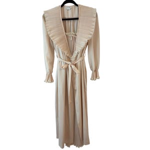 May include: A light beige vintage robe with a pleated collar and cuffs. The robe has long sleeves, a tie waist, and a delicate lace detail at the neckline. The robe is hanging on a black hanger.