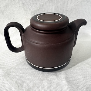Puede incluir: Una tetera de cerámica de color marrón oscuro con acabado mate, asa curva y pico corto. La tapa es circular con un centro marrón oscuro y un fino anillo blanco. Un fino anillo blanco a juego rodea la base del cuerpo de la tetera.