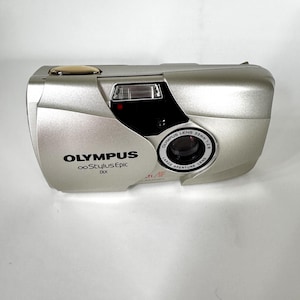 May include: A silver Olympus Stylus Epic DLX 35mm film camera featuring a 35mm 1:2.8 lens, a built-in flash, and a gold-coloured shutter button. The front body displays the Olympus logo and the Stylus Epic DLX model name in black text.