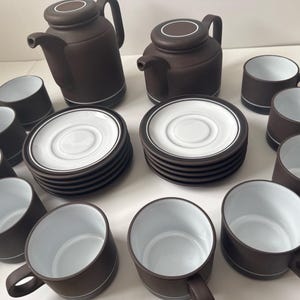 Puede incluir: Un juego de té de cerámica marrón con tazas y platillos interiores blancos. El juego incluye dos teteras, múltiples tazas con asas y varios platillos. Cada pieza tiene una línea de acento blanco cerca de la base y el borde.