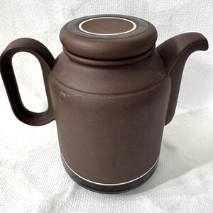 Puede incluir: Una tetera de cerámica marrón mate con cuerpo cilíndrico, asa curva y pico vertedor. La tapa presenta un diseño circular con un acento de anillo blanco. Una fina línea blanca rodea la base de la tetera, que tiene una banda inferior de color marrón oscuro.