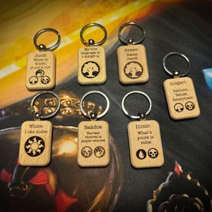 Könnte beinhalten: Holz-Schlüsselanhänger mit silbernen Schlüsselringen. Jeder Schlüsselanhänger hat ein einzigartiges Design und Text, darunter Sätze wie "Jund it out", "Ramp ranch" und "I like dudes". Die Schlüsselanhänger sind wahrscheinlich auf ein Spiel oder Fandom ausgerichtet.