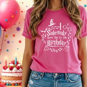 Puede incluir: Una camiseta rosa con el texto blanco "I Solemnly Swear that it's My Birthday". El diseño incluye un sombrero de mago, gafas y un pastel. La camiseta se combina con pantalones cortos de mezclilla. Globos y una tarta de cumpleaños se ven en el fondo.