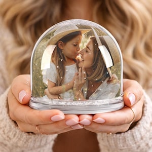 May include: A clear snow globe with a silver base, held in a person's hands. Inside, a photograph shows a woman and a child wearing straw hats, embracing. The background is blurred, suggesting an outdoor setting. The globe is filled with glitter.