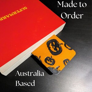 May include: A red book with the word "SUPERMARK" in yellow text. A Halloween-themed bookmark with orange background and black pumpkin faces is inserted into the book. The text "Made to Order" and "Australia Based" are also visible.
