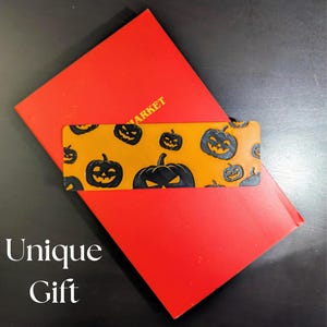 May include: A rectangular orange and black Halloween-themed bookmark with a repeating pattern of jack-o'-lanterns, resting on a red rectangular card. The words "Unique Gift" are displayed in white text.