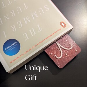 May include: A red and white bookmark with a star and swirl design is inserted into a book titled "The Summer I Turned Pretty." The book has a Penguin logo and text that reads "From the bestselling author of To All The Boys I've Loved Before." The words "Unique Gift" are at the bottom.