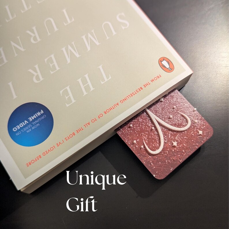 May include: A red and white bookmark with a star and swirl design is inserted into a book titled "The Summer I Turned Pretty." The book has a Penguin logo and text that reads "From the bestselling author of To All The Boys I've Loved Before." The words "Unique Gift" are at the bottom.