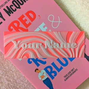 Puede incluir: Una etiqueta rectangular con un patrón ondulado en tonos rosa y blanco, con el texto "Your Name" en blanco. La etiqueta está colocada encima de un libro rosa con las palabras "RED" y "BLUE" visibles.