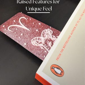 May include: A red bookmark with white raised features, including a heart and floral design, is placed in a book. The book has a Penguin logo and the text "FROM THE BESTSELLING AUTHOR OF TO ALL THE BOYS". The bookmark has the text "Raised Features for Unique Feel".