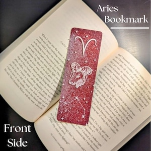 May include: A red Aries bookmark with a white ram illustration and constellation design. The bookmark is rectangular and is shown next to an open book. Text on the bookmark reads "Aries Bookmark".
