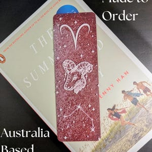 May include: A burgundy bookmark featuring the Aries zodiac symbol and constellation, with the text "Made to Order" and "Australia Based". The bookmark is placed on top of a book.