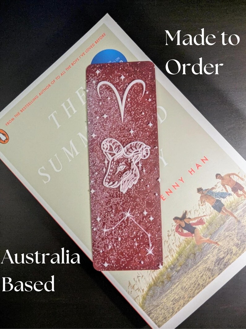 May include: A burgundy bookmark featuring the Aries zodiac symbol and constellation, with the text "Made to Order" and "Australia Based". The bookmark is placed on top of a book.