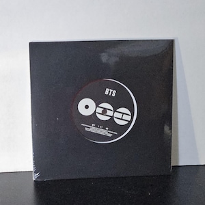 May include: A square black vinyl record sleeve encased in clear plastic wrap. The center label is circular and black with white text reading BTS and a graphic logo consisting of three white shapes, including a circle, a rectangle, and a horizontal bar.