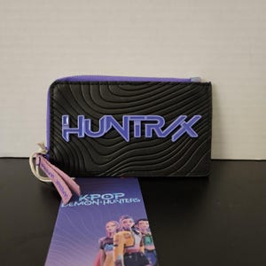May include: Black wallet with a purple zipper and the word "HUNTRAX" in blue. A tag with the words "K-POP DEMON HUNTERS" and a graphic of three people is attached to the wallet.