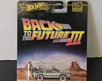 May include: A Hot Wheels Premium die-cast model of the Back to the Future III time machine. The silver car has red-rimmed wheels and is packaged on a black card with the movie title and logo. The card also features the Hot Wheels and Pop Culture logos.
