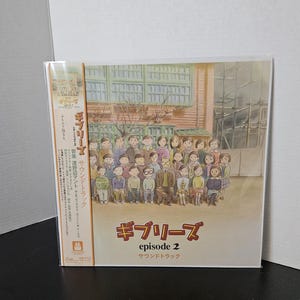 May include: A sealed CD case featuring the artwork for "Ghiblies Episode 2" soundtrack. The cover art depicts a group of people in front of a building. The title is written in Japanese and English. The CD is in a clear plastic sleeve.