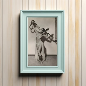 May include: A framed black and white photograph of a woman with a snake. The woman is wearing a decorative outfit and holding the snake above her head. The frame is a light blue color and is hanging on a light wood-paneled wall.