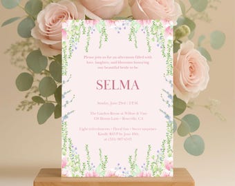 以下が含まれることがあります： ピンク色の「SELMA」という名前が書かれた結婚式の招待状で、花のイラストで囲まれています。招待状にはイベントの詳細が含まれており、木製のスタンドに表示されています。テキストは「私たちの美しい花嫁を称え、愛と笑いと花で満たされた午後を過ごしましょう」と書かれています。