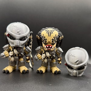 May include: Three Predator figurines. Two complete figures with detailed armour, skull accents, and articulated limbs. The third is a detached mask. The figures are primarily grey, tan, and black, with red accents. One figure has a keychain.