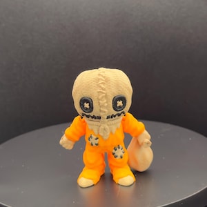May include: A small, orange figurine of a character from the horror film "Trick 'r Treat." The figure has a burlap sack head with black button eyes and a stitched mouth. It wears an orange jumpsuit and holds a beige sack.