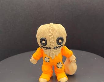 May include: A small, orange figurine of a character from the horror film "Trick 'r Treat." The figure has a burlap sack head with black button eyes and a stitched mouth. It wears an orange jumpsuit and holds a beige sack.