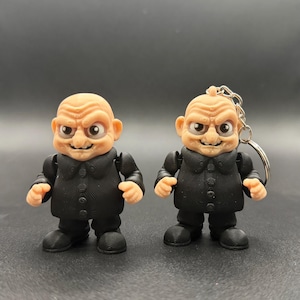 May include: Two small, articulated figurines of a bald, stern-faced character in a black suit. One figure has a keychain attached. The figures have a light skin tone and are posed against a dark background. The figures are likely toys or collectibles.