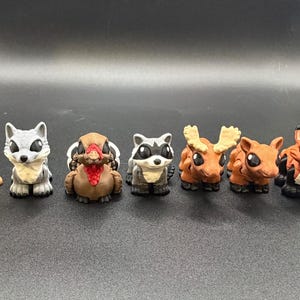 May include: A collection of seven miniature animal figurines, including an otter, wolf, turkey, raccoon, moose, and fox. The figurines are hand-painted with detailed features and stand on a dark surface.
