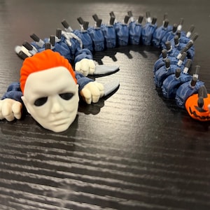 May include: A multi-segmented articulated figure featuring a white mask with orange hair at the front, followed by a long blue body with multiple small grey knives protruding from each segment, ending in a small orange pumpkin-like head.