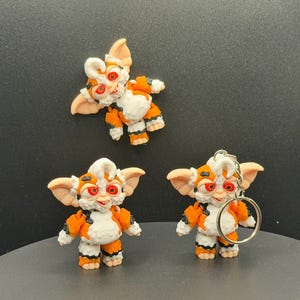 May include: Three handmade figurines of a creature with orange, white, and black markings. One figurine is a keychain. The creatures have large ears and red eyes. The figurines are on a dark surface.