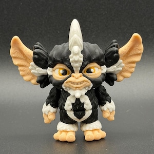May include: A small, three-dimensional printed figurine of a creature with large, pointed ears, a central white crest, and a textured black, white, and peach-colored body. The figure has wide, amber-colored eyes and a small, detailed facial expression.
