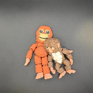 May include: Two articulated monkey figurines. One is orange with a smiling face and the other is brown with a white chest. Both have jointed limbs and are posed together on a dark gray surface.