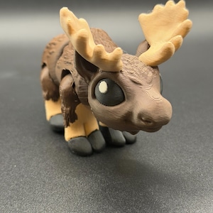 May include: A 3D-printed moose figurine in shades of brown, tan, and black. The moose has large, expressive eyes and detailed antlers. The figurine is standing on a dark surface.