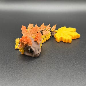 May include: A brown articulated dragon figurine featuring a series of orange and yellow leaf-shaped scales along its back and head, ending in a large, bright yellow maple leaf-shaped tail segment, with large black eyes and a smooth matte finish.
