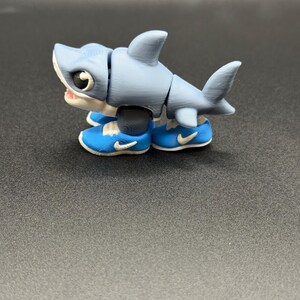 May include: A 3D printed articulated shark figurine in light blue with a white underbelly and large black eyes. The shark is wearing bright blue sneakers with white soles and a white swoosh logo on the sides. The body has visible layer lines.