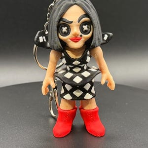 May include: A 3D-printed keychain featuring a doll-like figure with black hair and a black and white diamond-patterned dress. The figure has red lips and red boots. The keychain is attached to a silver metal ring.