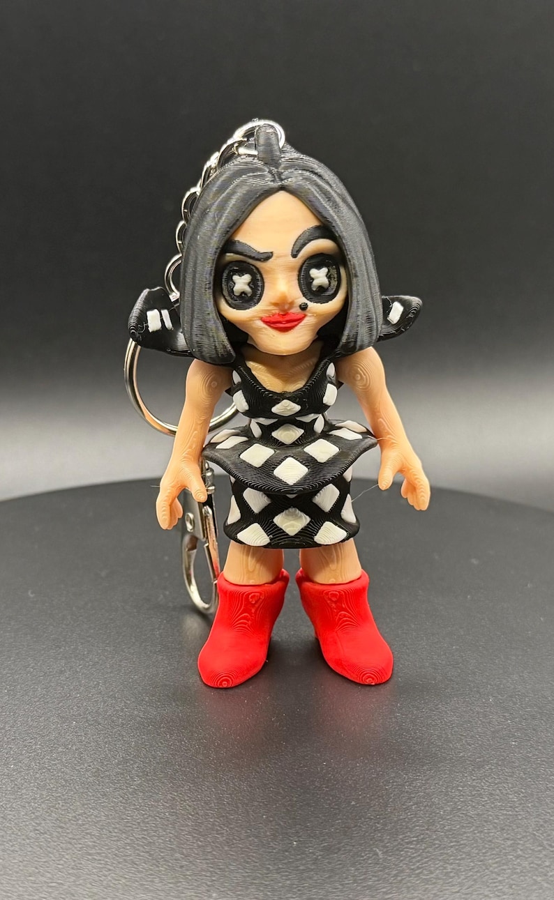 May include: A 3D-printed keychain featuring a doll-like figure with black hair and a black and white diamond-patterned dress. The figure has red lips and red boots. The keychain is attached to a silver metal ring.