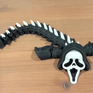May include: A black and white articulated figure featuring a ghost-like mask with a long, segmented, spine-like tail. The mask is white with black eye and mouth cutouts, framed by a black hood. The tail consists of repeating black segments with white spikes.