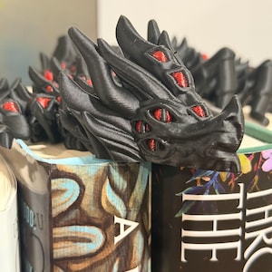 May include: A black dragon ornament with red eyes sits on a pile of books. The dragon is intricately designed, with textured scales and a fierce expression. The books have colourful covers, contrasting with the dark dragon.