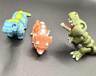 May include: Three colorful 3D-printed dinosaur toys. A blue and yellow dinosaur is on the left, an orange and white dinosaur in the middle, and a green dinosaur with an open mouth on the right. The toys are on a dark surface.