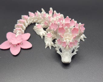 May include: A white and pink dragon ornament with a floral design. The dragon is decorated with pink flowers and has a matching pink flower beside it. The ornament is on a dark grey surface.
