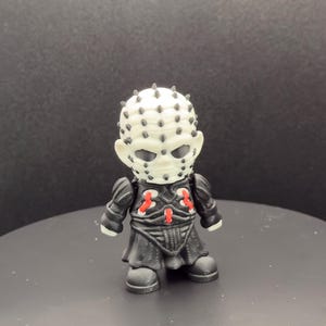 May include: A small, stylized figurine of a character with a white head covered in black pins and dark eye sockets. The figure wears a black outfit with red accents. The figurine is standing on a dark surface.