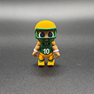 May include: A 3D-printed figurine of a football player. The figure wears a yellow helmet and uniform with green accents. The number "10" is printed on the chest. The figure has a cartoonish design with large eyes and white wristbands.