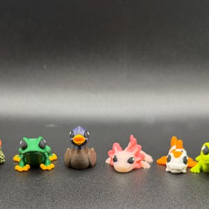 May include: A collection of six small animal figurines including a green turtle with an orange shell, a green frog with orange feet, a brown duck, a pink axolotl, a white and orange koi fish, and a light green frog, all arranged in a single row.