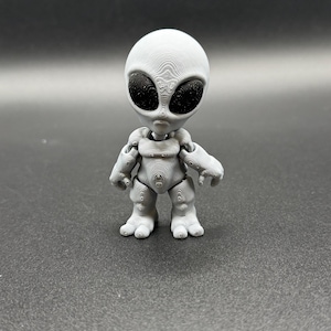 May include: A grey 3D-printed alien figure with large black eyes and a detailed, textured surface. The alien has a large head, small arms, and stubby legs, standing upright against a dark background. A unique decorative item.