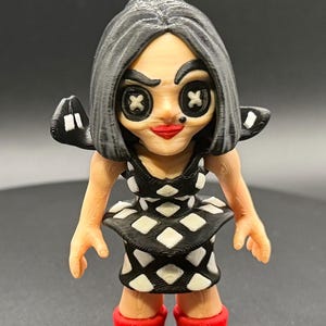 May include: A 3D-printed figurine of a character with button eyes and a red mouth. The figure wears a black and white diamond-patterned dress and red boots. The hair is gray, and the figure has small wings.