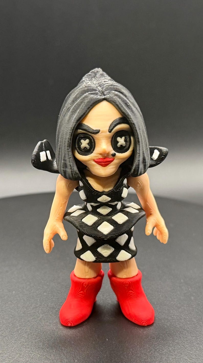 May include: A 3D-printed figurine of a character with button eyes and a red mouth. The figure wears a black and white diamond-patterned dress and red boots. The hair is gray, and the figure has small wings.