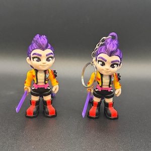 May include: Two figurines of a character with purple hair, yellow jacket, black shorts, and red and black boots. Each figure holds a purple sword. One figure has a keychain attached. The figures are on a dark gray surface.