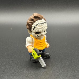 May include: A small figurine of a character wearing a textured white mask with black stitching details, a bright yellow apron over a white shirt, blue pants, and black boots. The character holds a lime green and silver toy chainsaw in one hand.